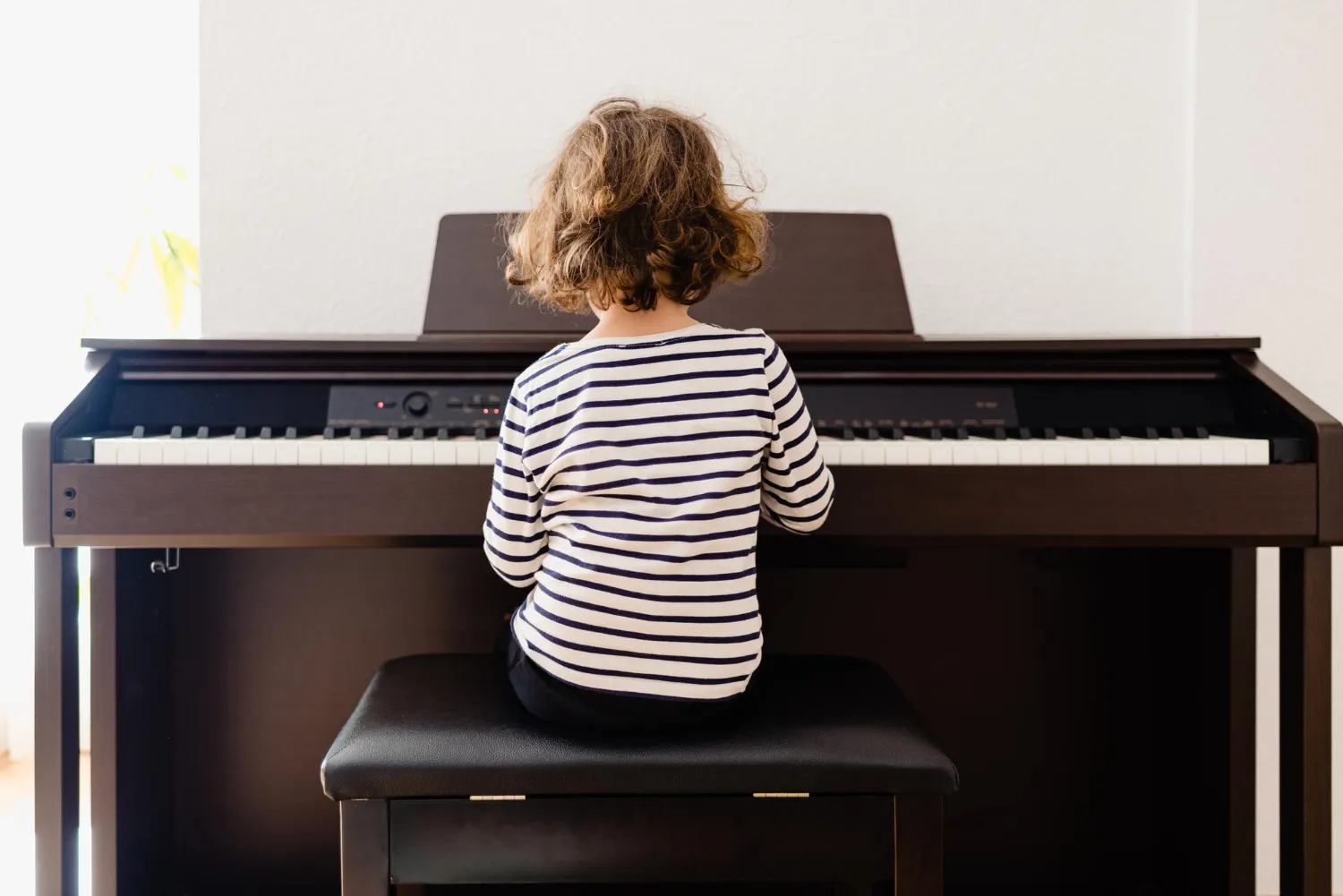Criança sentada tocando piano digital, vista de costas, com camiseta listrada.